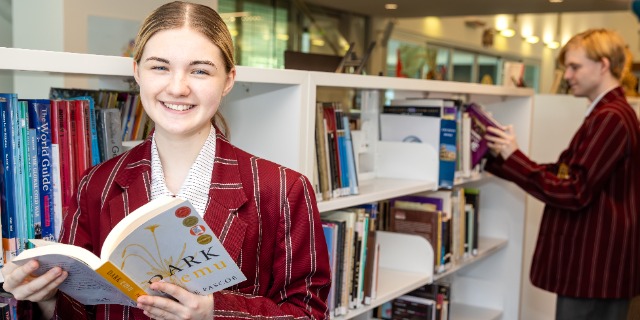 A student is in the library. She is facing towards the camera, smiling and holding a book in her hand. There is a student behind her, choosing a book from the shelf.